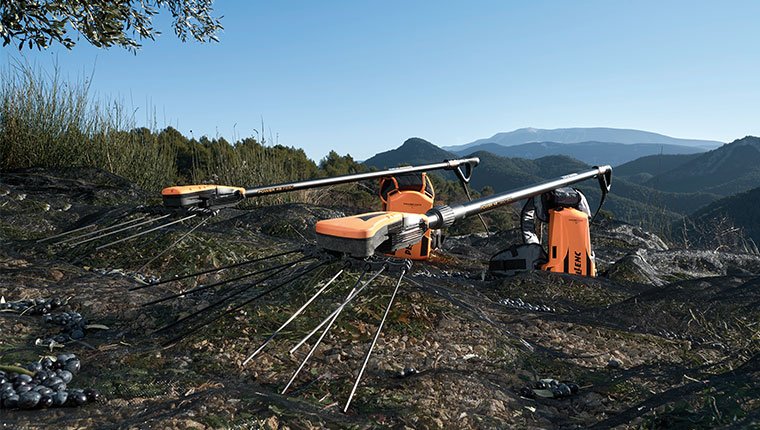 Pellenc telescopic olive and nut shaker tools with harvesting heads and battery system positioned in a mountain orchard